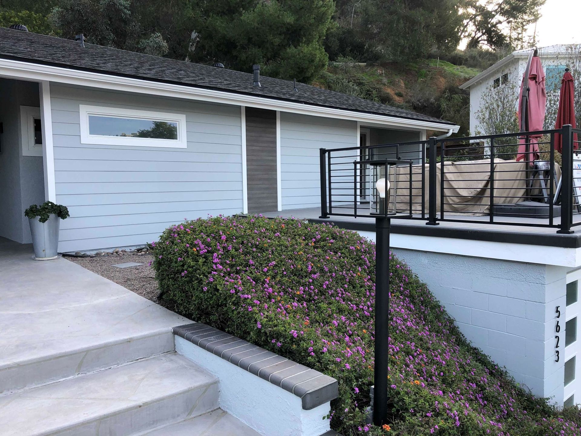 Light blue house with dark roof, small porch, and steps leading to entrance. Lush greenery in front.