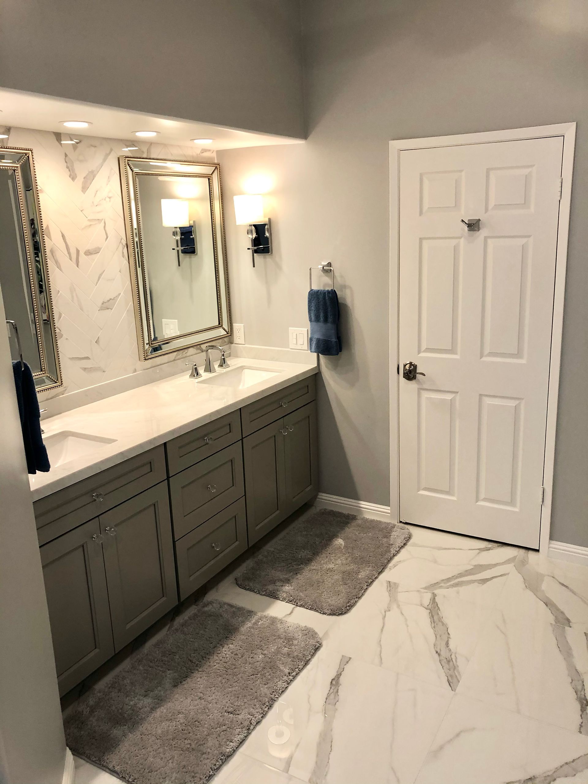 Bathroom with double vanity, marble-look floor, gray cabinets, and a white door.