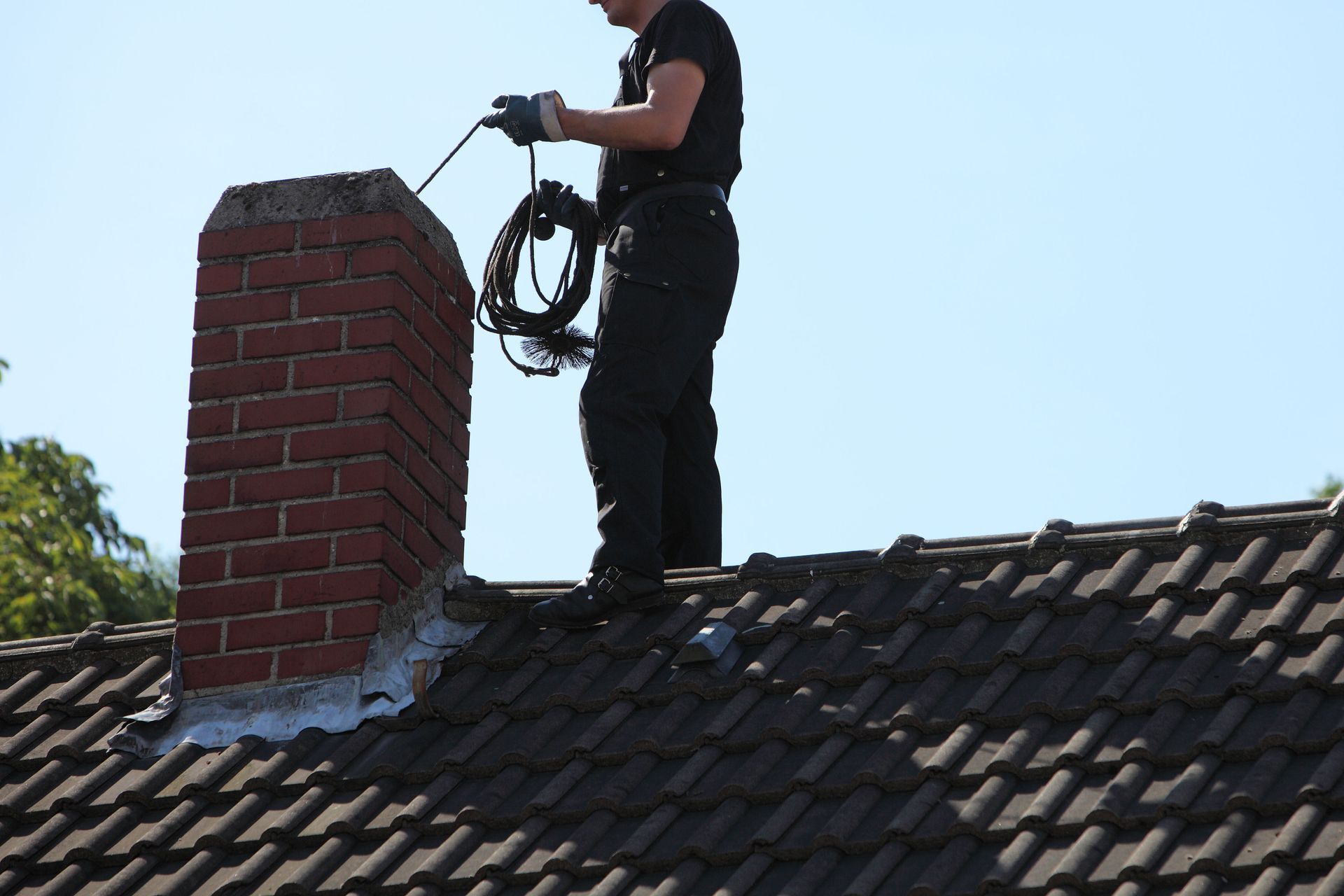 Person on a roof cleaning a brick chimney. Holding a tool, wearing black clothing, and a bright blue sky is in the background.