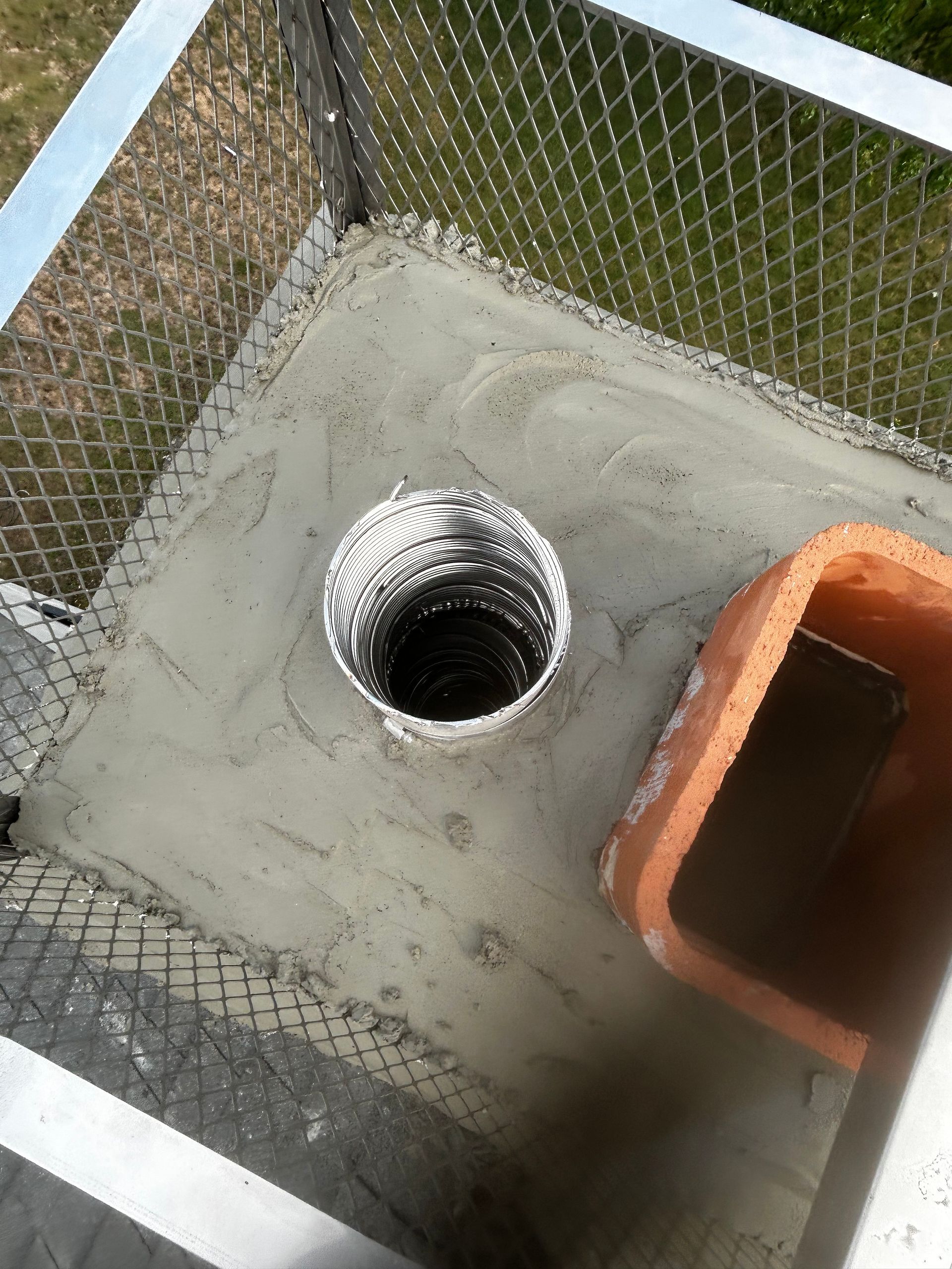 Concrete utility pad with a round metal opening beside a chain-link fence and an orange plastic bin