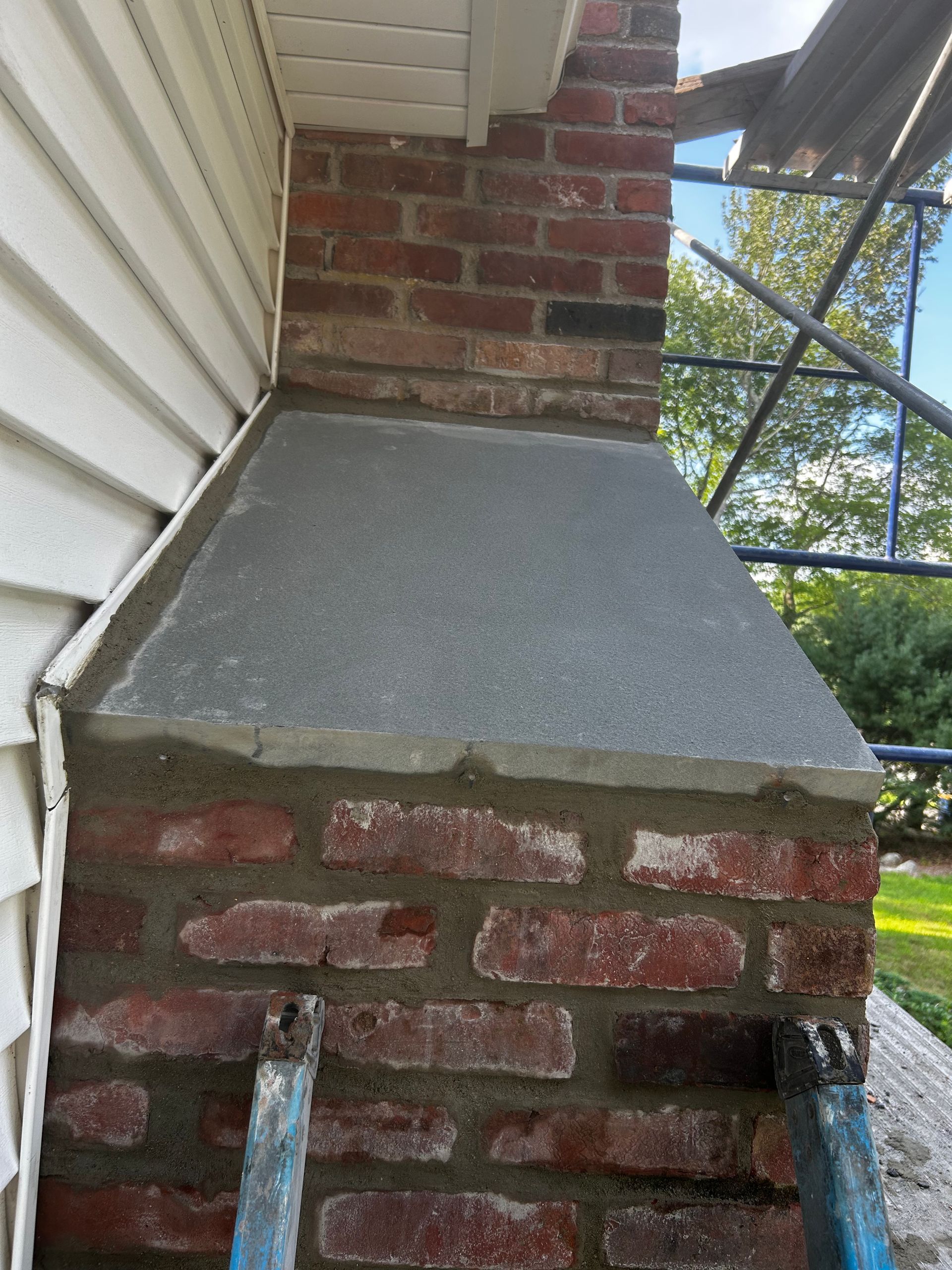 Brick porch support pier with a gray concrete cap beside white siding and a brick chimney