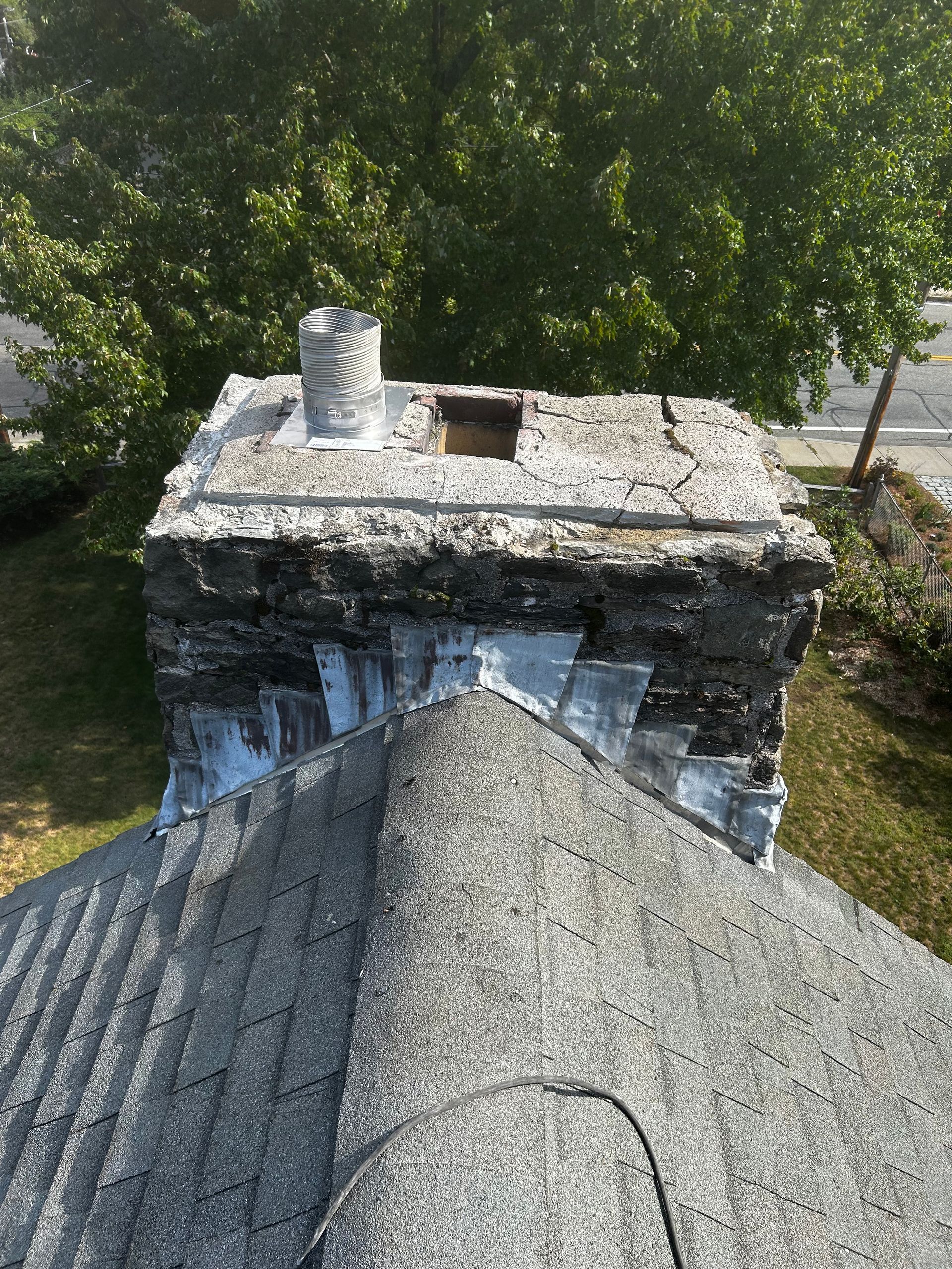 Damaged brick chimney with missing top bricks beside a gray shingled roof and green trees behind
