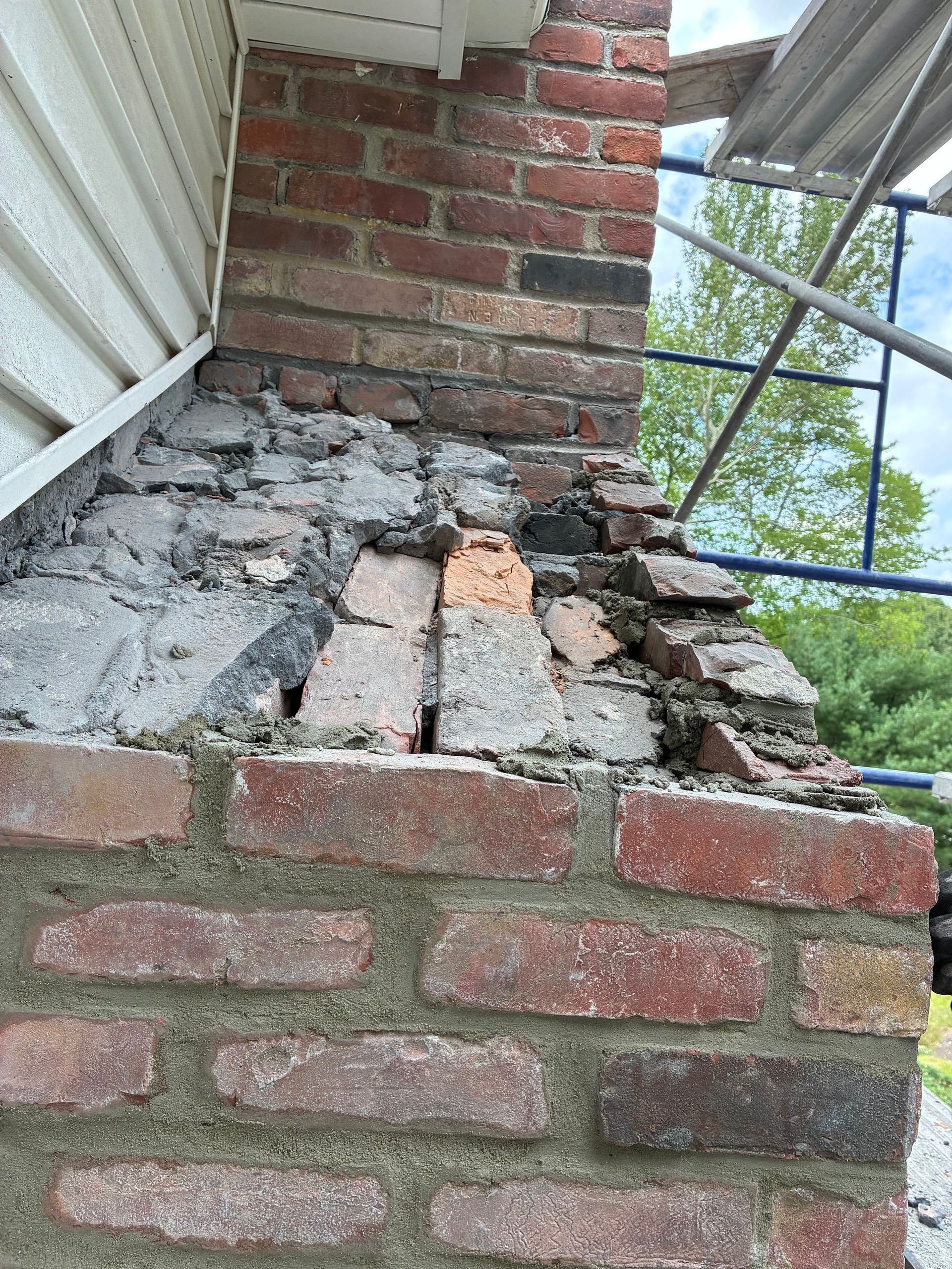 Damaged brick chimney with crumbling mortar beneath a roof overhang and metal scaffolding.