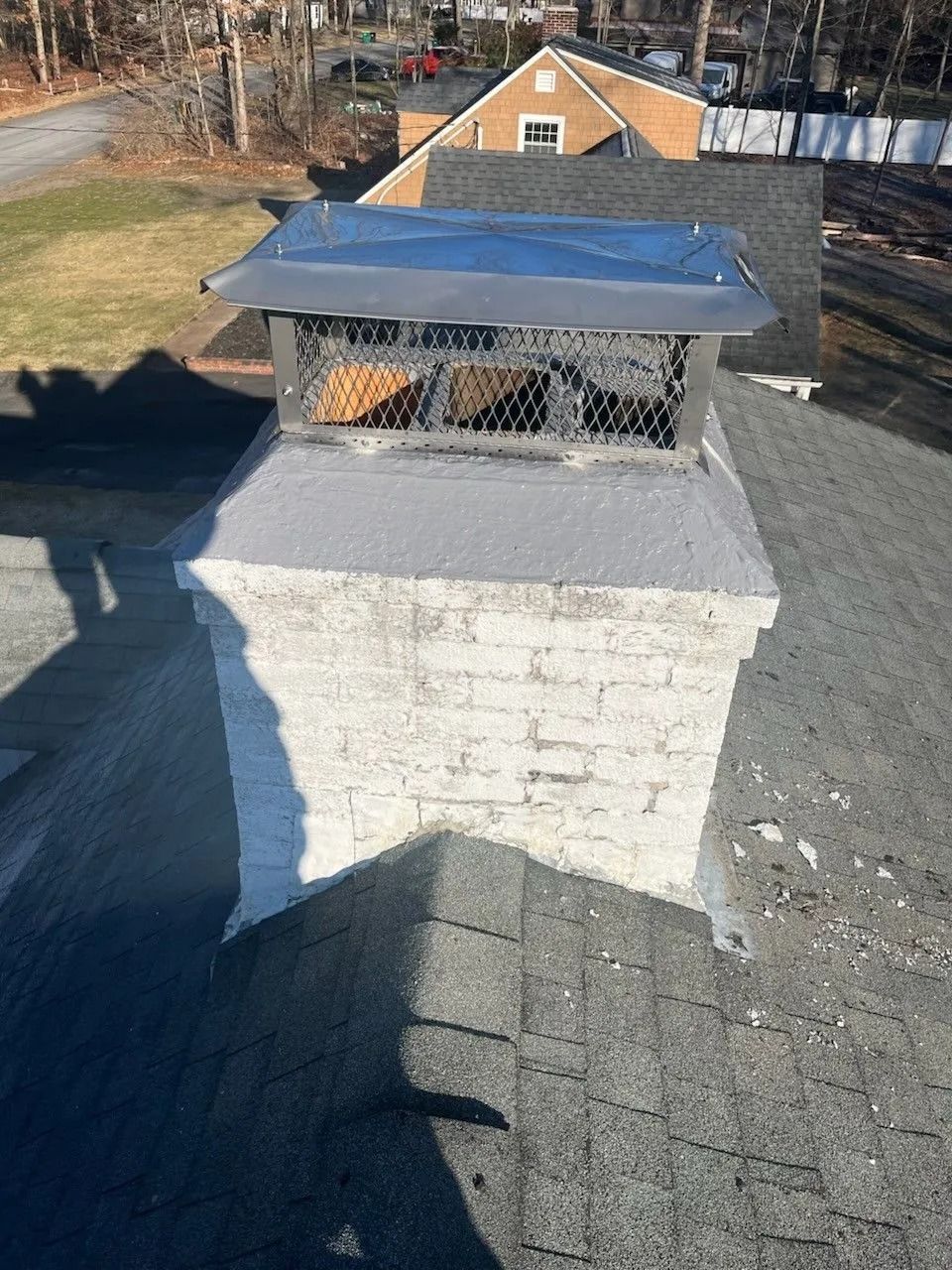Chimney with metal cap on a gray roof, in front of buildings and trees.