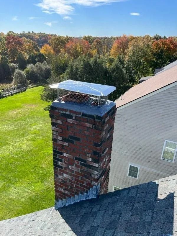 Brick chimney on a rooftop with a metal chimney cap, surrounded by fall foliage under a blue sky.