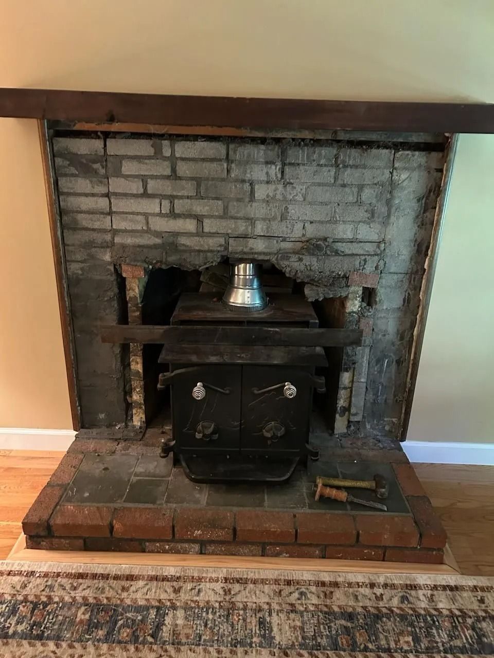 A wood-burning stove installed in a brick fireplace, surrounded by partially demolished brick and wooden frame.