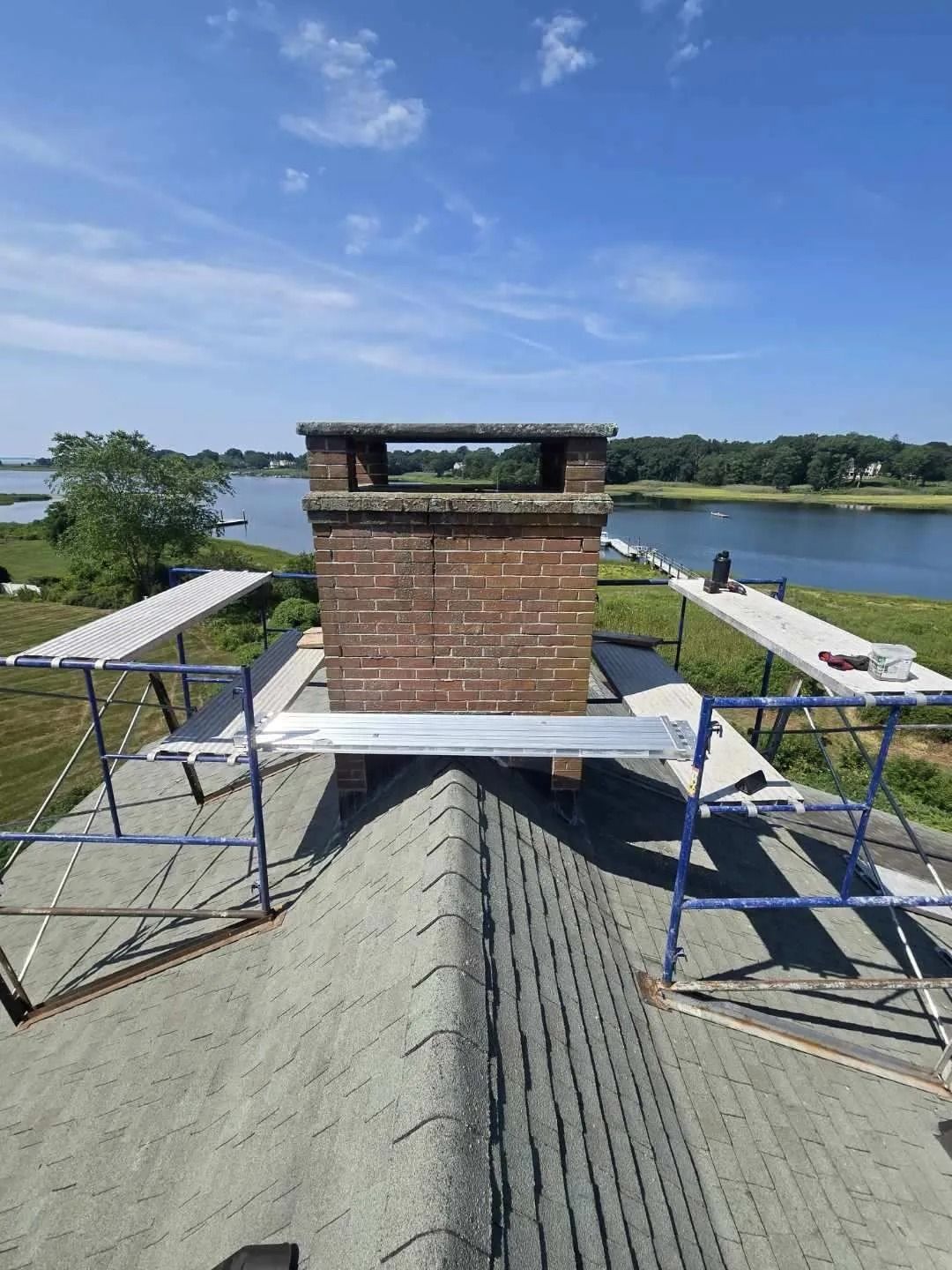 Chimney on rooftop with scaffolding, overlooking water and trees on a sunny day.
