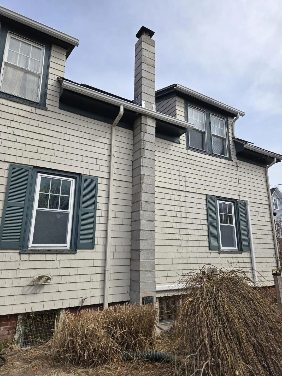 Side view of a light beige house with green shutters and a tall gray chimney. Brown shrubbery in foreground.