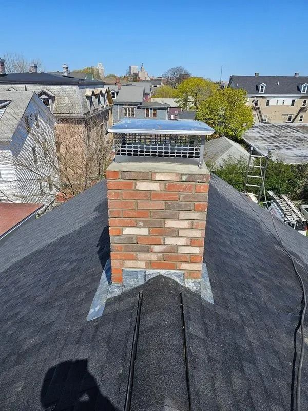 Brick chimney with metal cap on a dark roof, surrounded by city buildings under a blue sky.
