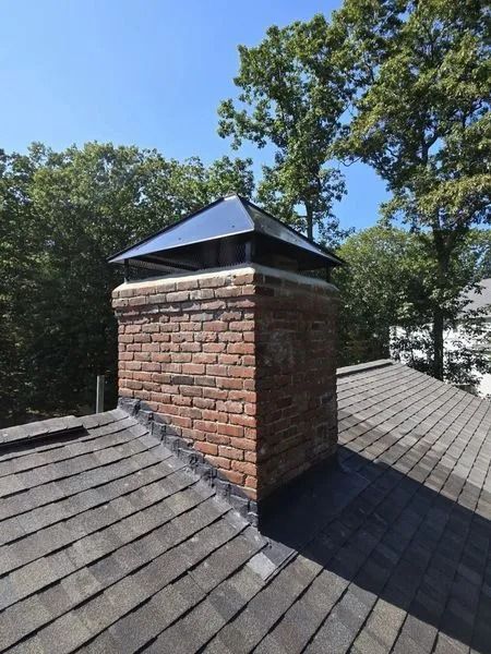 Brick chimney on a shingled roof with a metal cap, trees in the background, blue sky overhead.