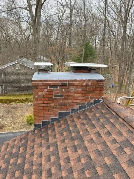Brick chimney with two metal caps on a brown shingled roof, trees in background.