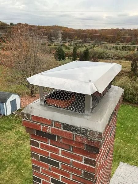 Chimney with a metal cap on a brick structure, against a background of trees and a cloudy sky.