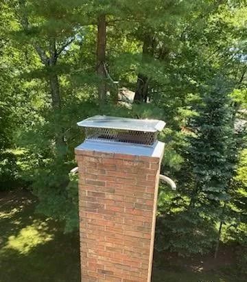 Brick chimney with a metal cap, surrounded by green trees.