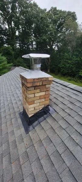 Brick chimney on a shingled roof with a metal cap. Trees are in the background.
