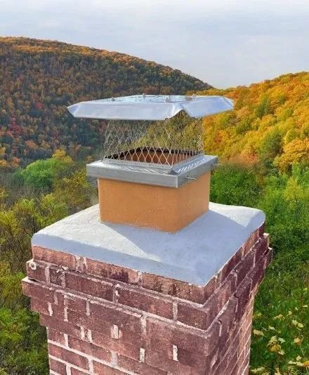 Chimney with metal cap and spark arrester, brick structure against a fall foliage background.