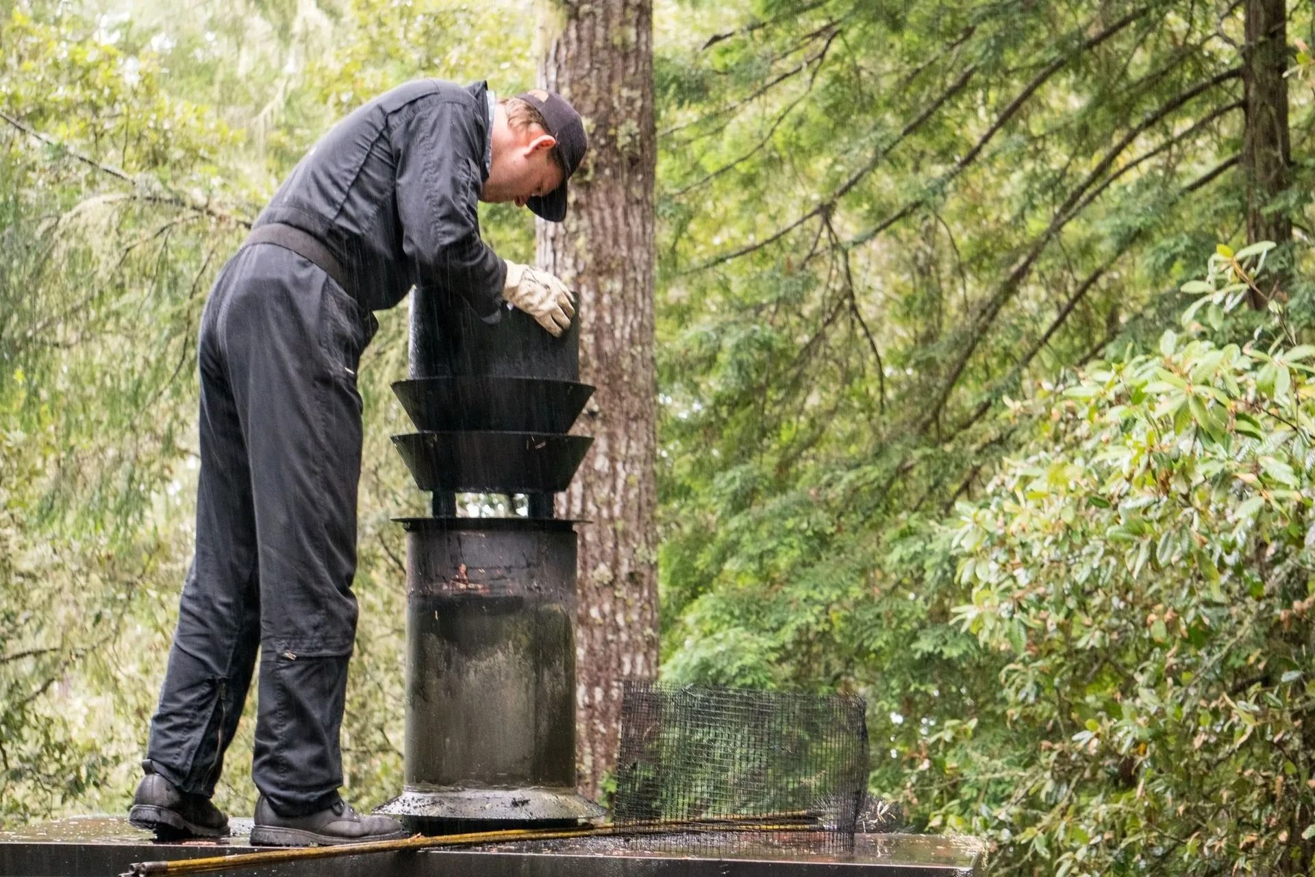 Man in dark coveralls inspecting a chimney cap in a wooded outdoor setting.