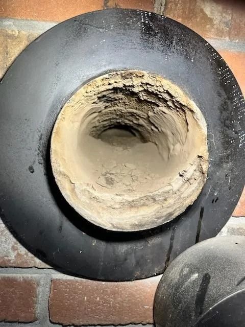 A chimney flue opening with significant creosote buildup, surrounded by a black metal collar, against a brick wall.