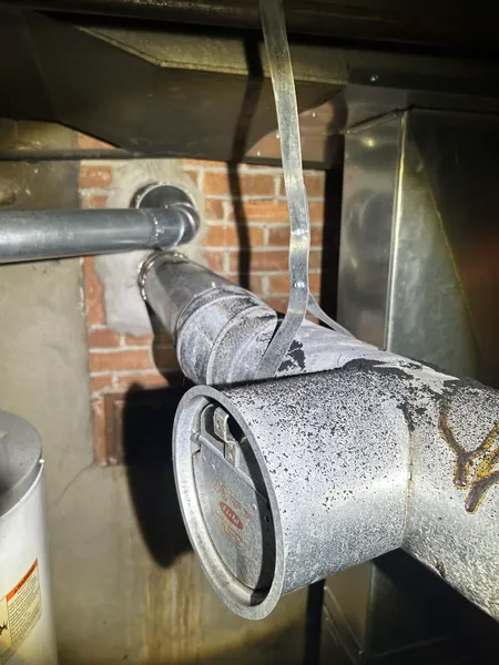 Metal furnace components connected to a brick wall with gray ductwork. A water heater is in the background.
