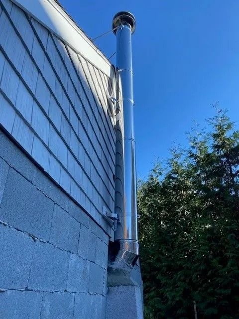 Silver chimney pipe attached to a gray, shingled house exterior. Blue sky and green trees in the background.