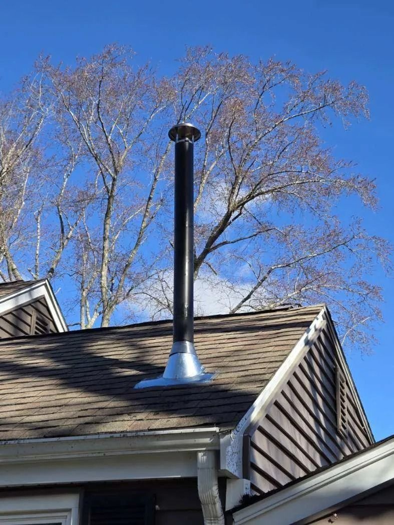 Black chimney on a brown roof, with a metal flashing and a blue sky with leafless tree branches in the background.