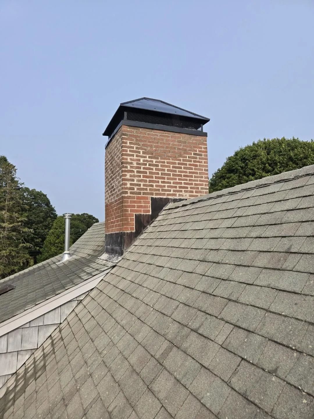 Brick chimney with a black cap on a gray shingle roof under a clear sky.