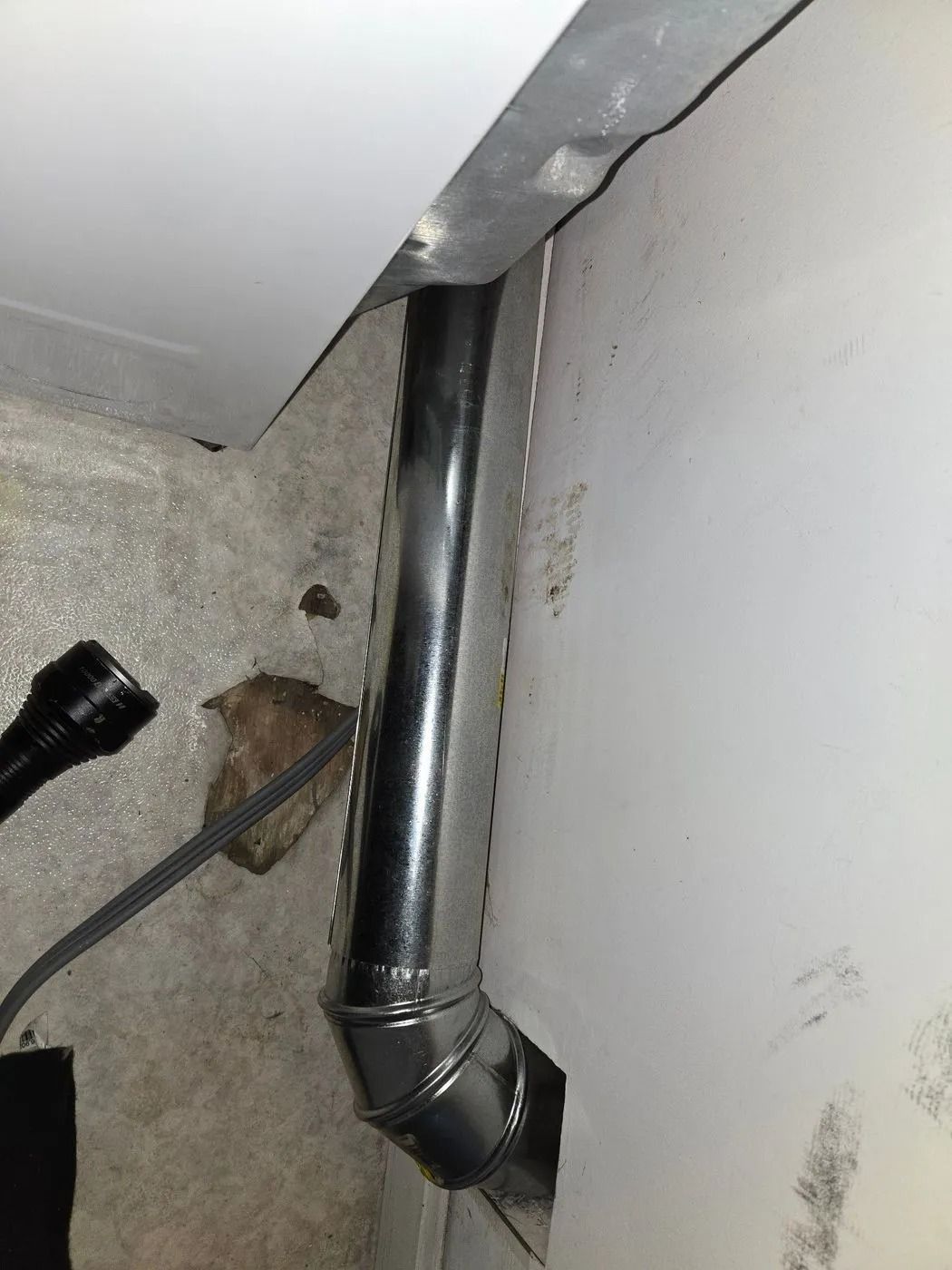 Metal dryer vent pipe exiting from wall, with a flashlight on the floor to the left.