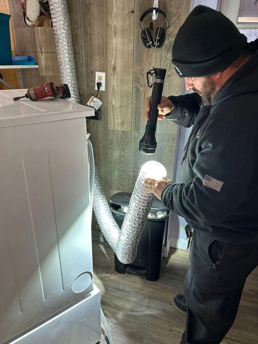 Man inspects dryer vent with flashlight, near a white appliance in a room with wood paneling.