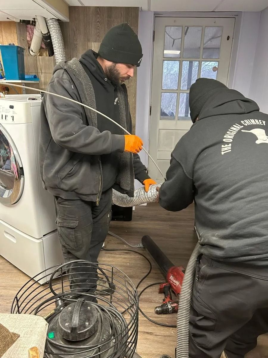 Two people cleaning a dryer vent. One holds a cleaning rod, the other operates a blower. Indoors, near a washing machine.