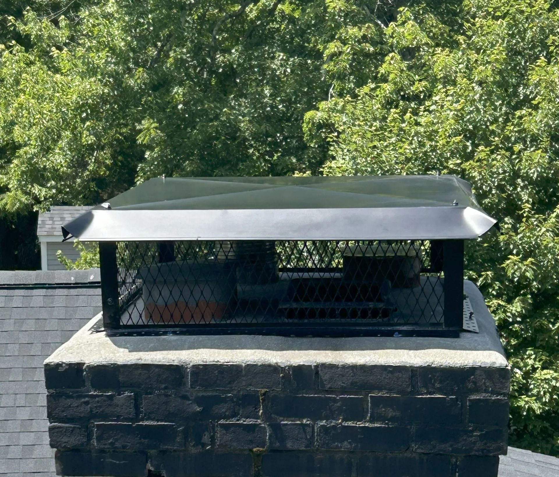 Chimney with black cap, screen, and concrete base against a backdrop of green trees.
