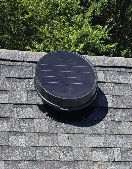 Black solar-powered roof vent on a gray shingled roof, trees in the background.
