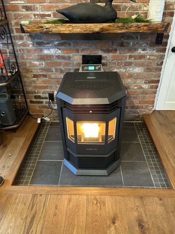 A pellet stove with a lit fire sits on tiled hearth in front of a brick wall and wooden shelf.