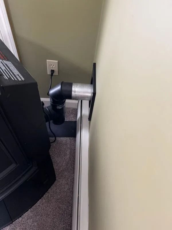 A black pellet stove exhaust pipe connected to a wall-mounted chimney. Beige wall, carpet.