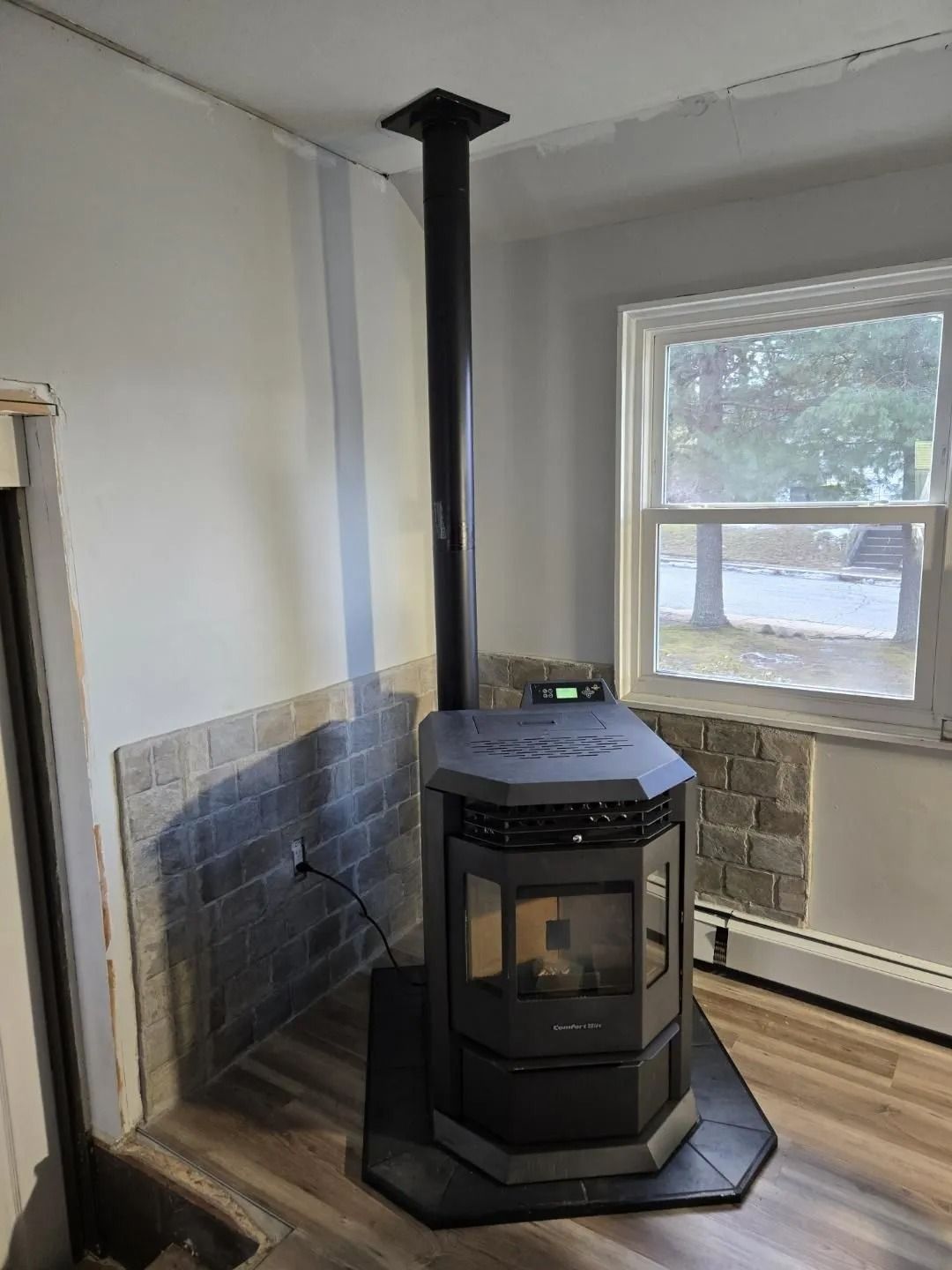 Pellet stove with black chimney pipe, installed against a partially tiled wall, next to a window.