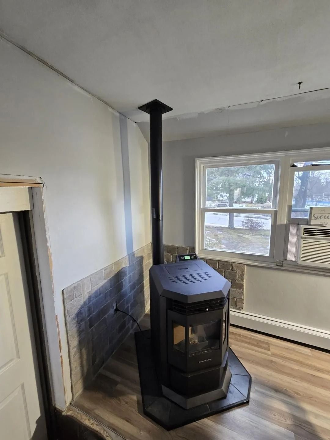 A dark-colored pellet stove with chimney in a room with a window, white walls, and wood flooring.