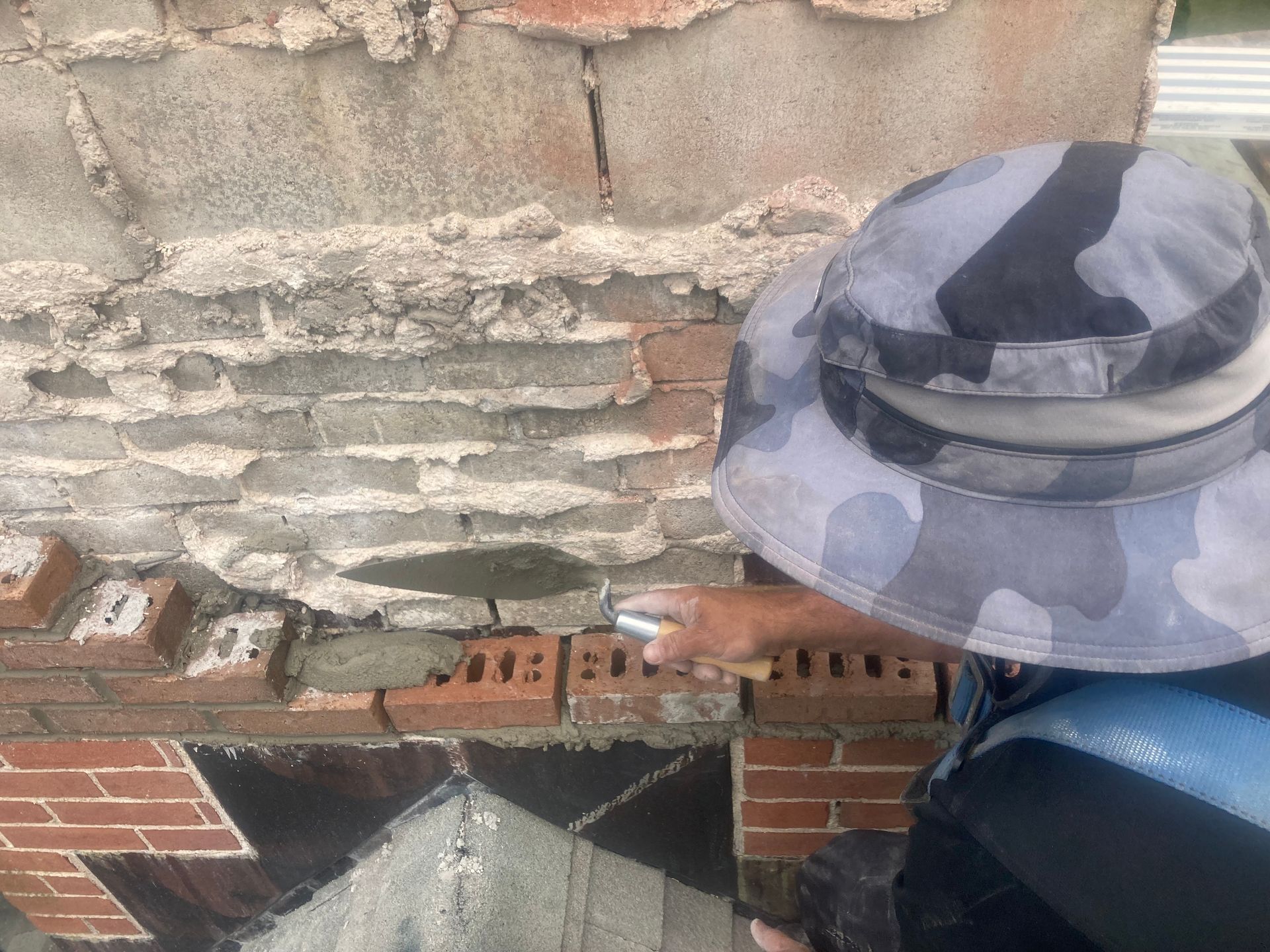 Person in camouflage hat pointing a trowel to brickwork while repairing a wall.