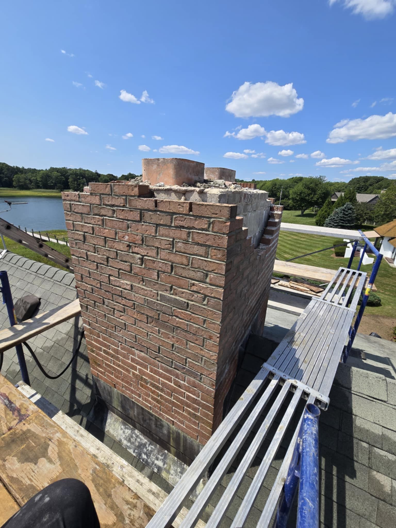 Brick chimney partially dismantled, scaffolding, blue sky, trees, and lake in background.