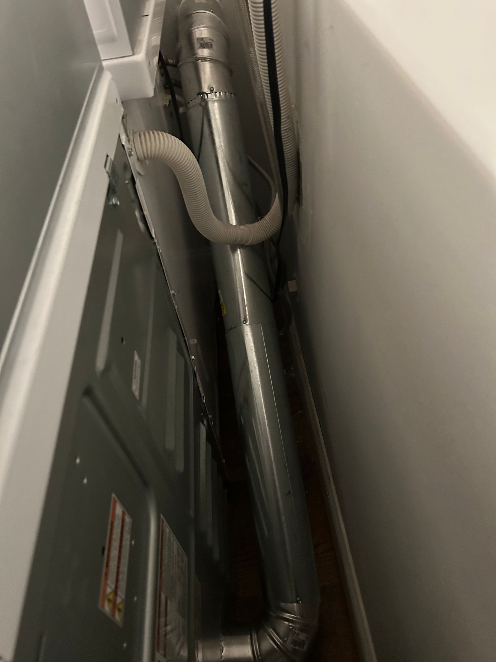 Inside view of a dryer and its metal exhaust vent. A flexible gray tube connects to the dryer's metal vent. The wall is white.