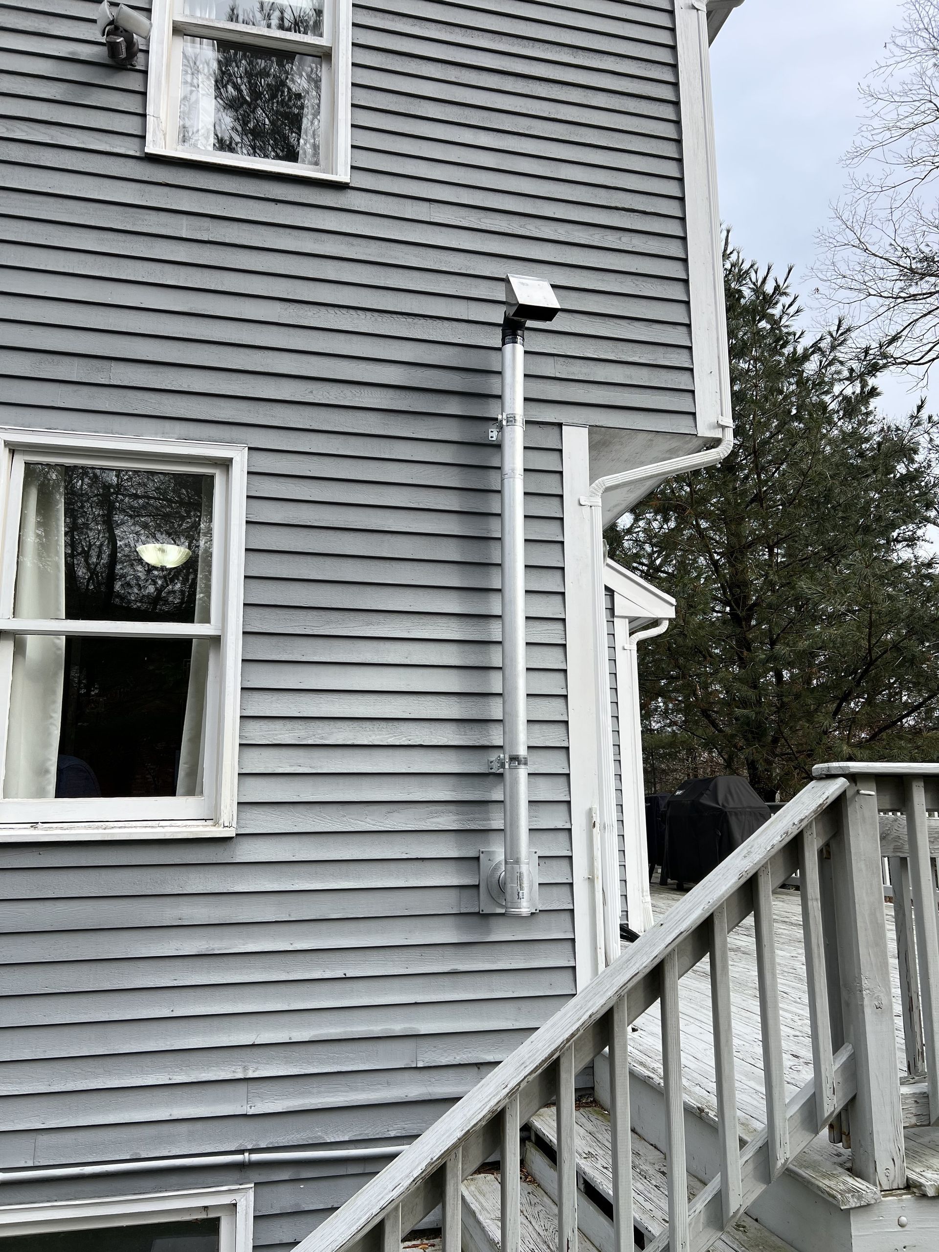 Gray clapboard house with white trim, windows, and exterior vent pipe. Wooden stairs and railing in foreground.
