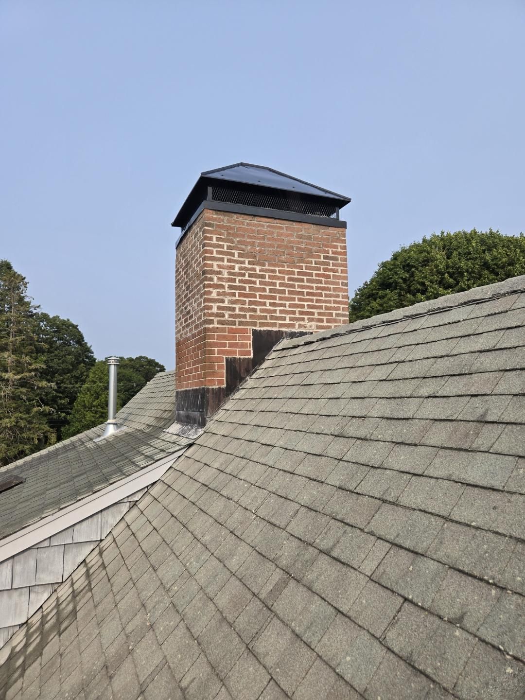 Brick chimney with a black cap on a gray shingled roof, trees in the background, blue sky.