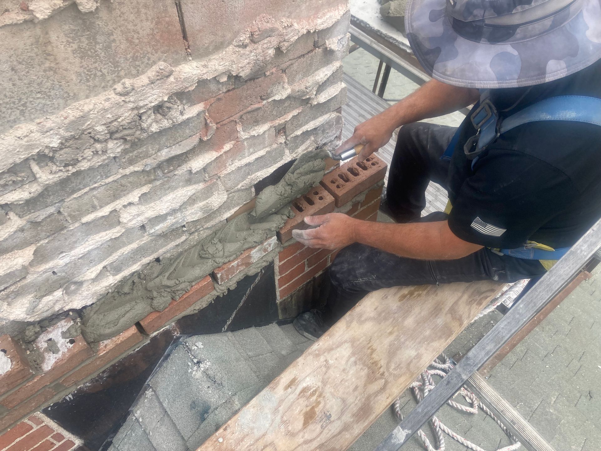 A person is bricklaying a chimney on a rooftop. Mortar and bricks are visible, with the person wearing a safety harness.
