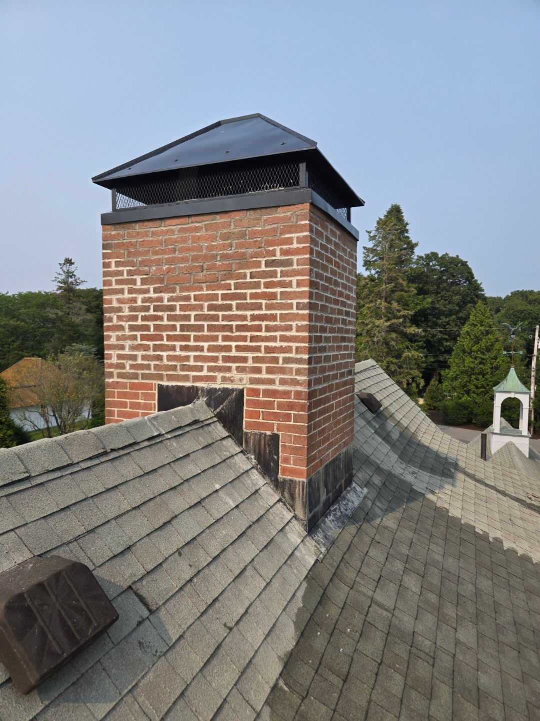 Brick chimney with a black cap on a gray shingled roof, trees in the background.