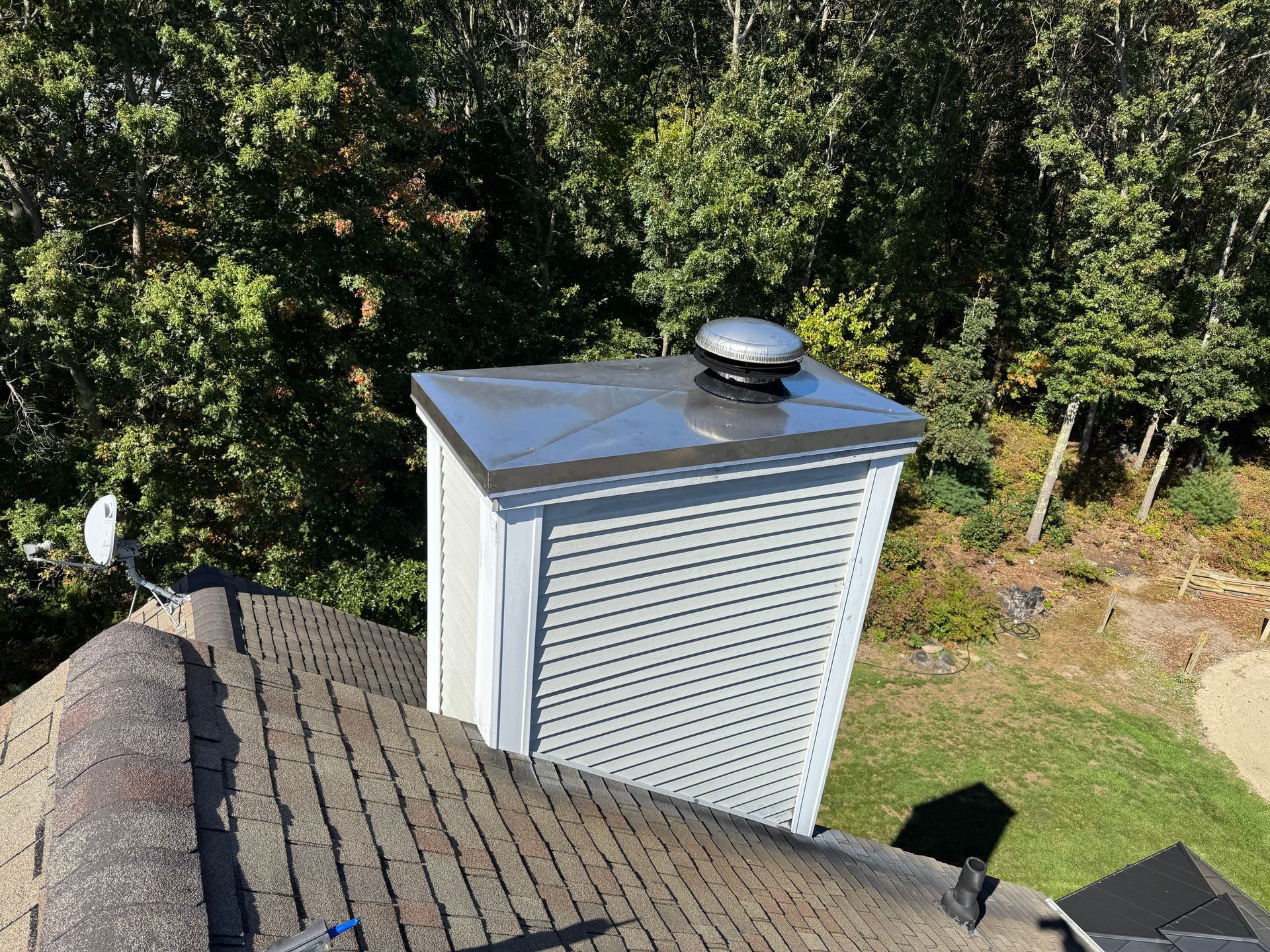Chimney on a roof with a metal cap and vent, surrounded by green trees.