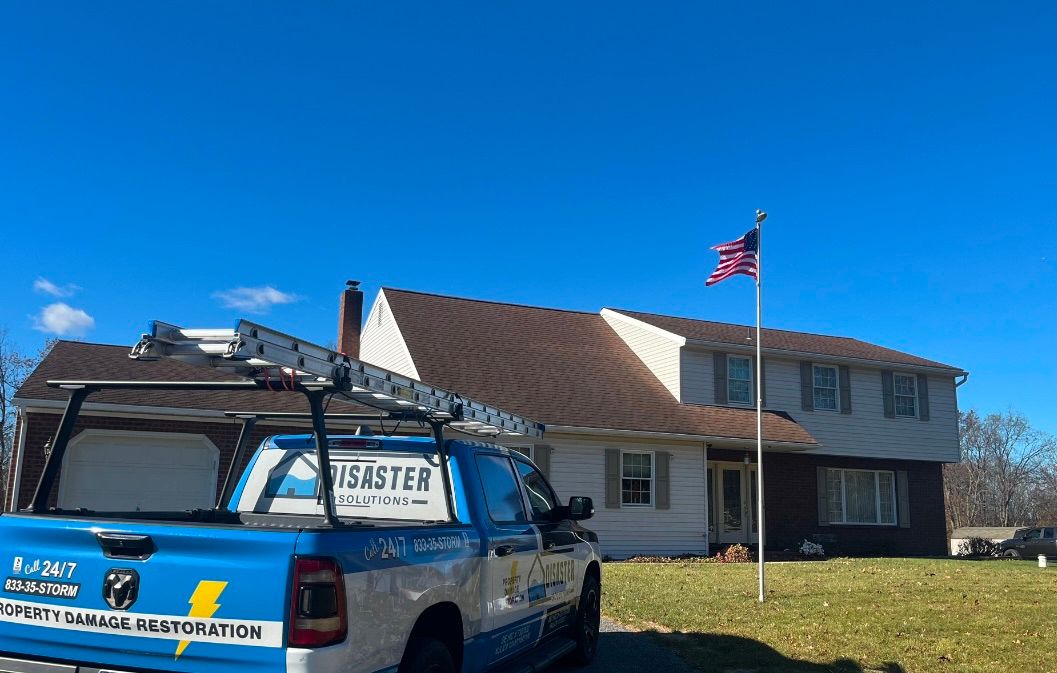 Blue truck with ladder parked in front of a two-story house with American flag. Clear sky.