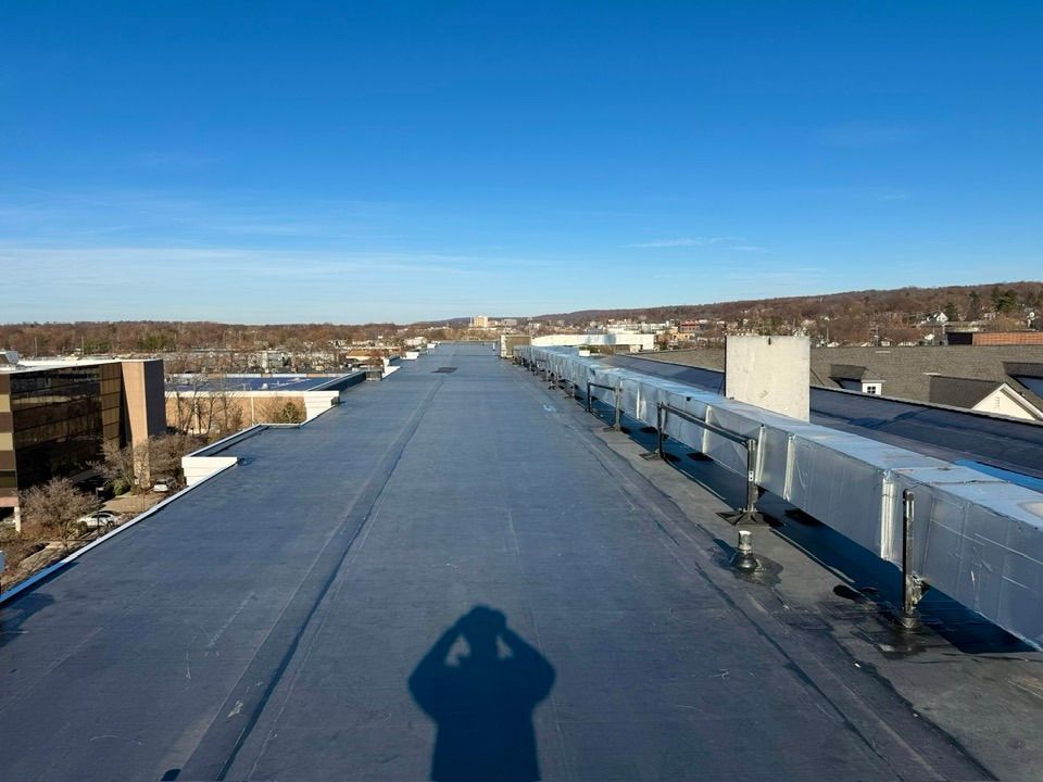 View from a rooftop on a sunny day; gray flat roof, long silver vent, trees, buildings, blue sky.