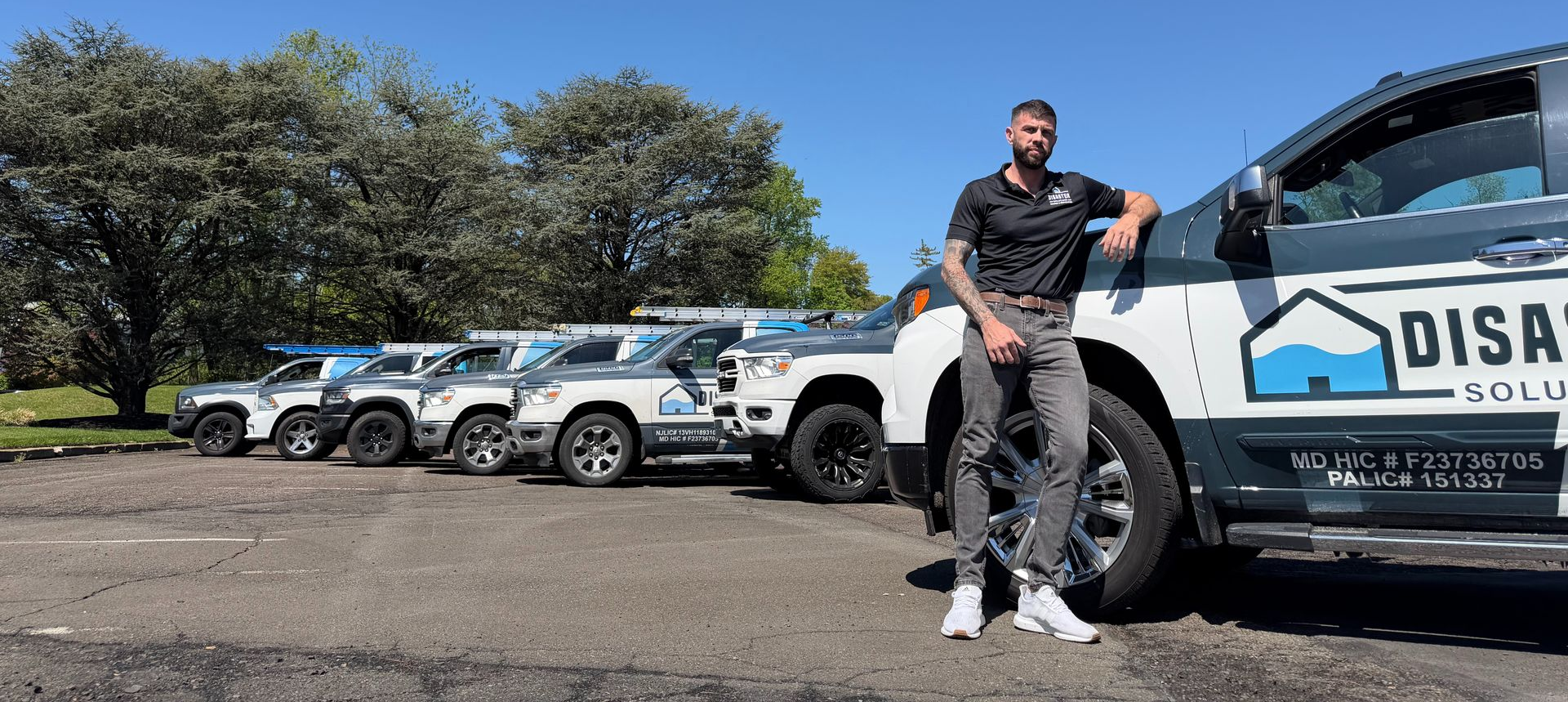 Man standing beside white off-road vehicles on a sunny gravel lot, with trees in the background.