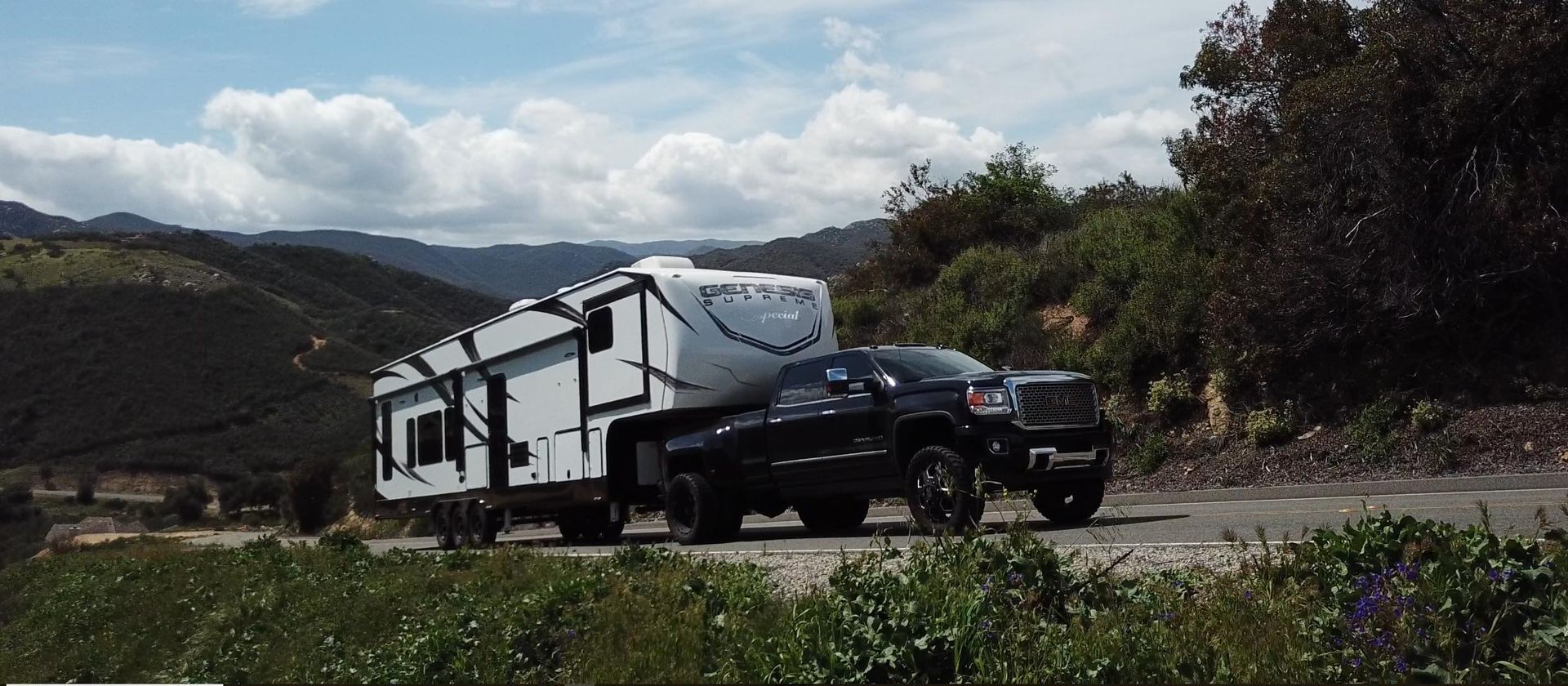 White RV parked on a mountain road with green hills and snowy peaks in the background.