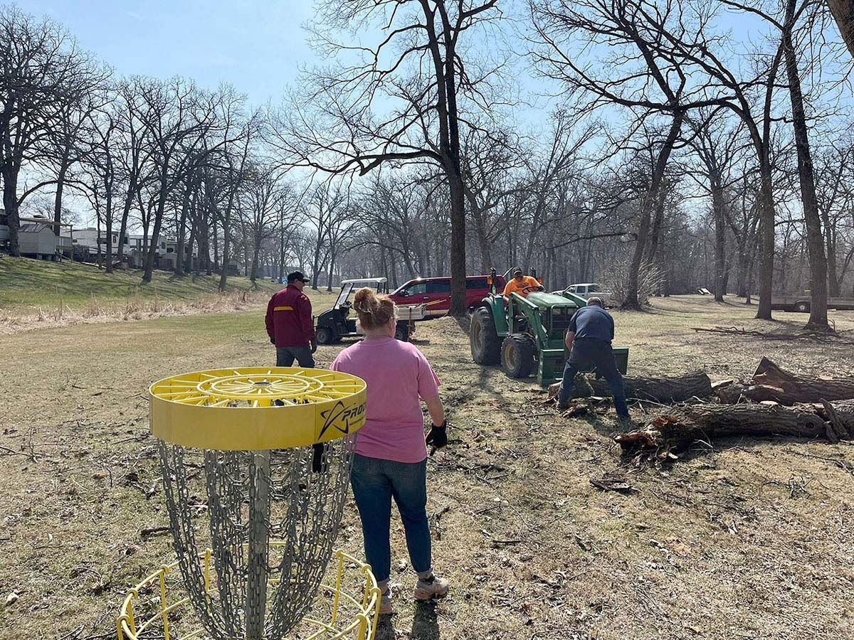 Disc golf basket in foreground; people clearing brush near a tractor in a park