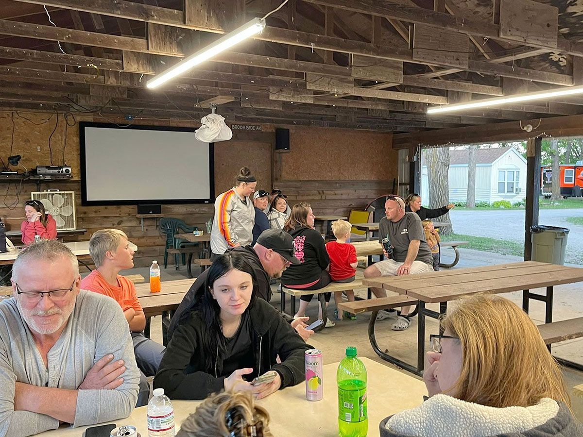 People gather at picnic tables under a pavilion with a projection screen