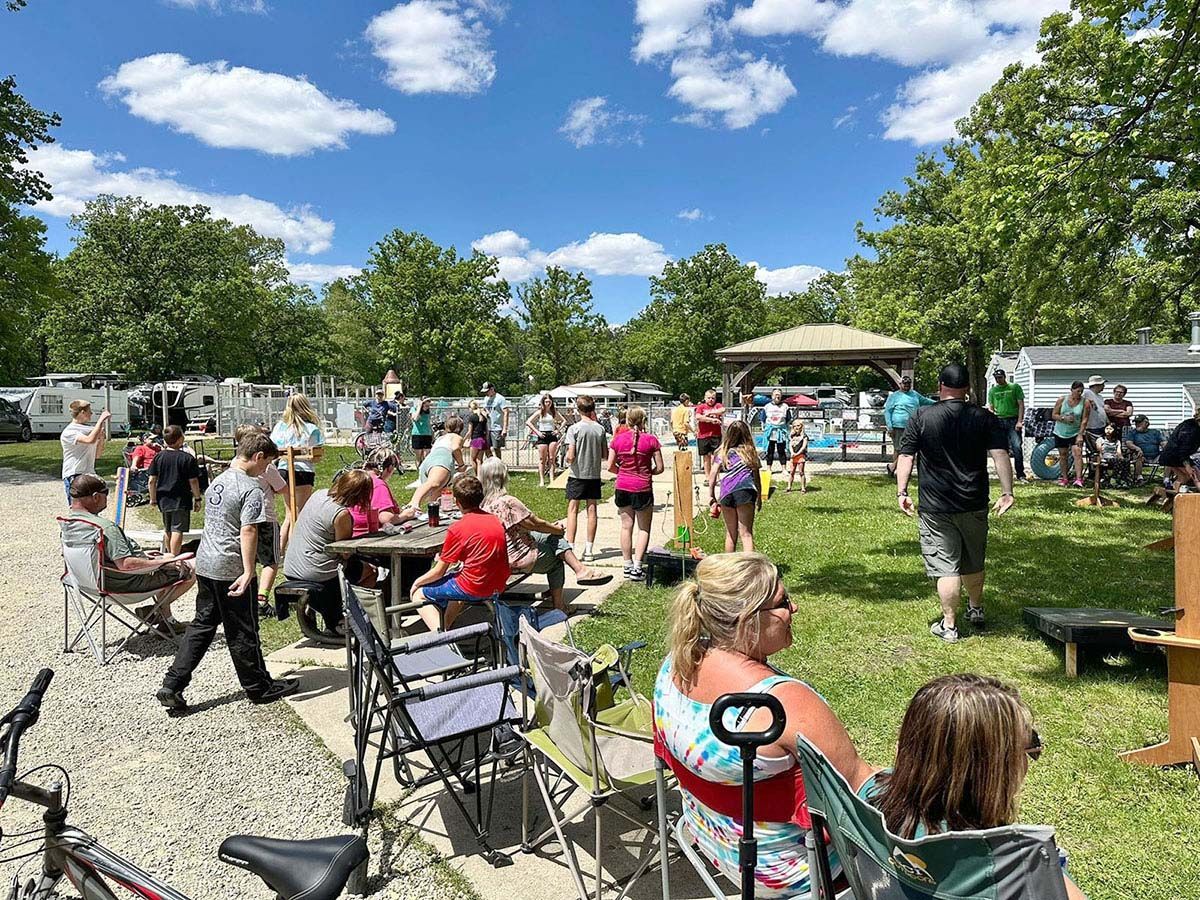 People gathered outdoors at a sunny park; some sitting in chairs, others standing