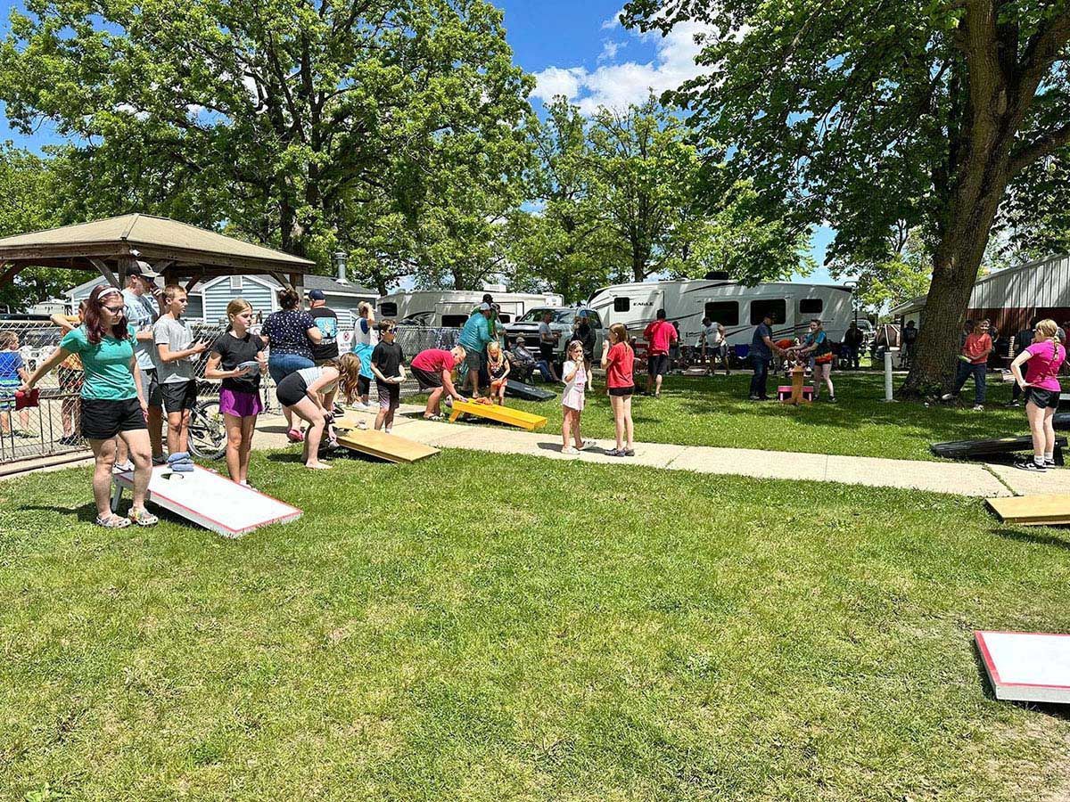 People playing cornhole on a grassy lawn with RVs in the background, under a bright blue sky
